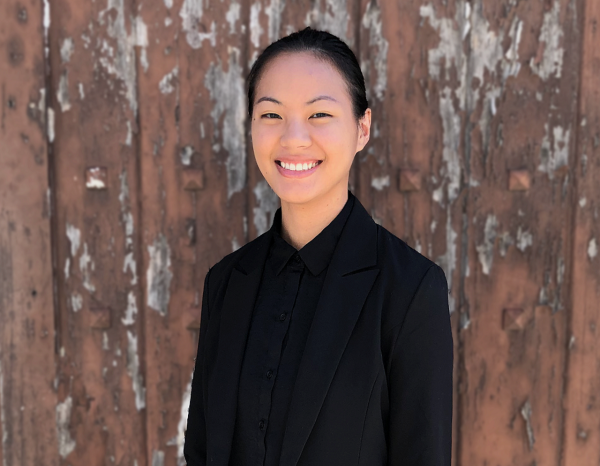 Xie Maggie Hill in all black smiling at the camera against a rustic wood background
