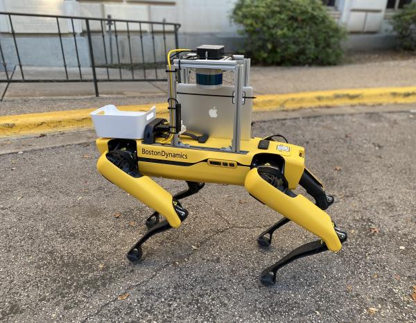 A yellow four-legged robot with an apple ipad attached stands on the sidewalk, poised to deliver cookies on campus