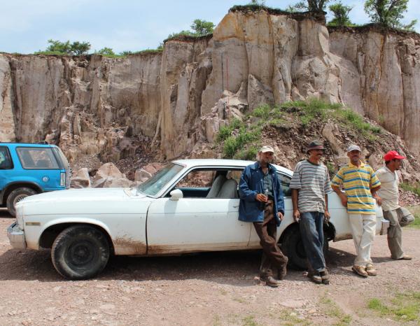 Four Mexican men in hats lean against a white sedan in front of a cliff of brown, rocky cantera stone