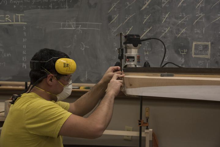 A student in yellow with headphones stoops down to look at a piece of wood he's working on in the Build Lab in front of a chalkboard