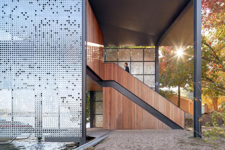 Exterior shot of a person ascending a wood paneled staircase with a reflective, almost transparent facade seen on the left and light filtering through trees on the right