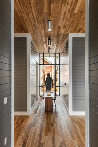 View looking down a hallway with dark walls with white trim on either side, a table in the middle with someone seen coming through the door. Grainy wood covers both the floor and ceilings