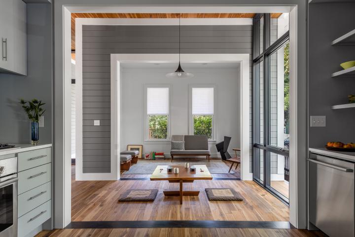 Shot taken from the kitchen looking through to the dining room in the middle and a living room on the far end. Walls are grey, horizontal paneling white white trim and the floor is heavily grained wood. The dining room table has a low profile close to the ground