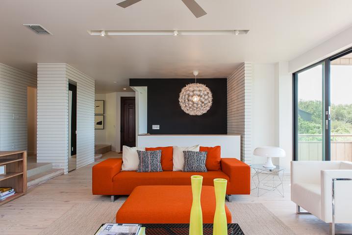 Interior shot of a living room with a bright orange couch, a black accent wall, a dramatic modern chandelier and natural light filtering in from a windowed door on the right