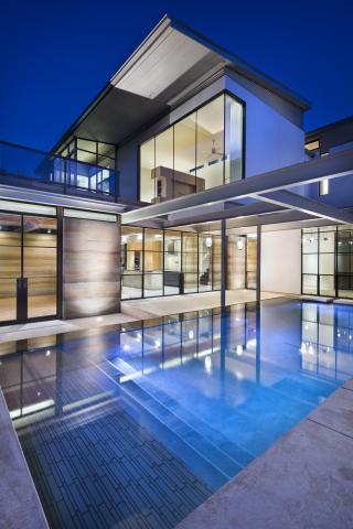 Night time shot of a backyard with a blue lit pool in the foreground and multiple levels of a modern looking, heavily windowed house in the background