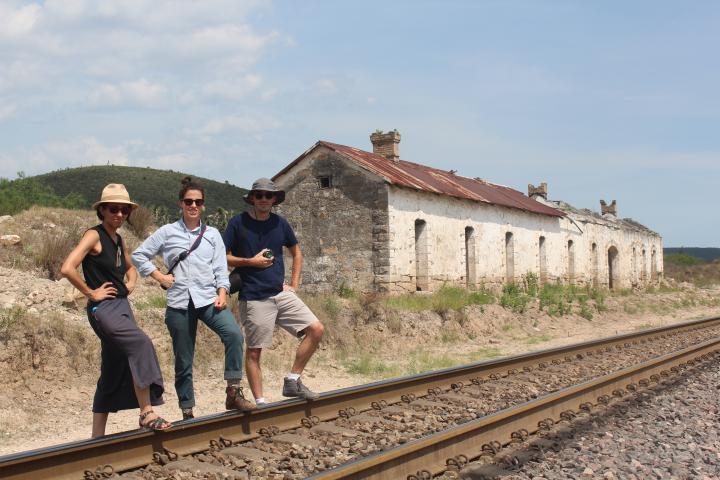 Three people stand along a train track in front of a historic stone building