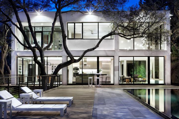 Exterior night shot of a backyard, with a glistening pool on the right, two white lounge chairs and a dramatic tree in front of a well-lit house