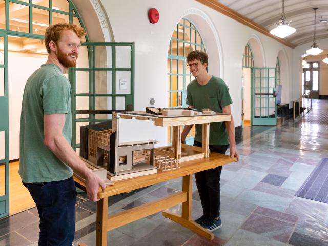 Two boys in green carrying a table with a large architecture model on it