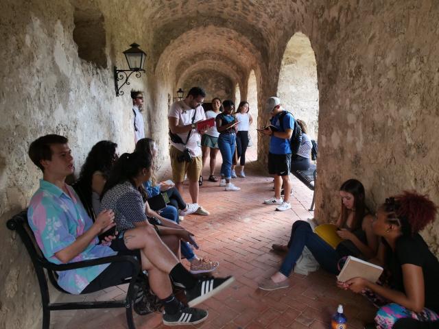 Students sitting with notebooks in a stone hallway with arched entrances