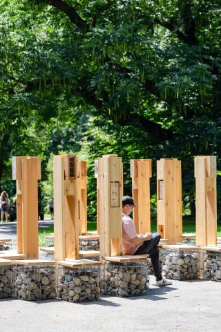 Photograph of a modular wood installation in a forested area of the Brooklyn Botanic Garden, with seating at the base of each module and built-in pollinator habitats at the top.