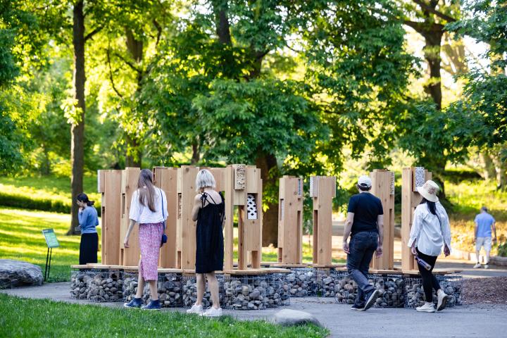 Photograph of a modular wood installation in a forested area of the Brooklyn Botanic Garden, with seating at the base of each module and built-in pollinator habitats at the top.