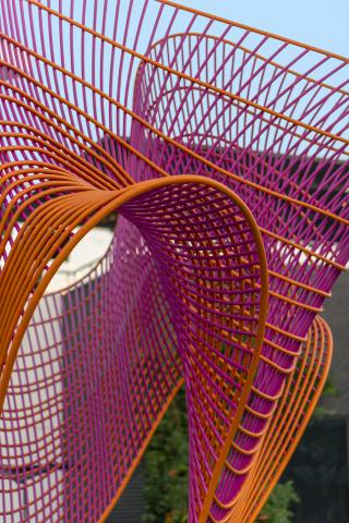 Large-scale public sculpture of interwoven magenta and orange powder-coated steel rods forming a dynamic, undulating woven form on a concrete plinth, set in a park at The Village Dallas. Photography by Wage Griffith