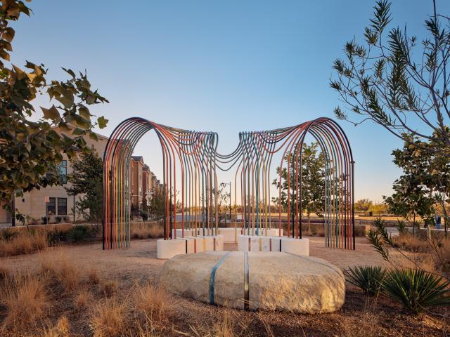 Detail view of a curving public sculpture featuring smooth white concrete segments joined by multicolored metal rods in pink, gold, teal, bronze, and silver, casting striped shadows on sandy ground. Photography by Casey Dunn